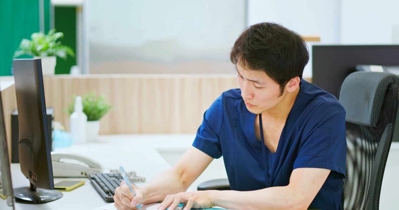 Male nurse in navy blue scrubs sitting at a desk, focused on writing notes with medical equipment nearby.