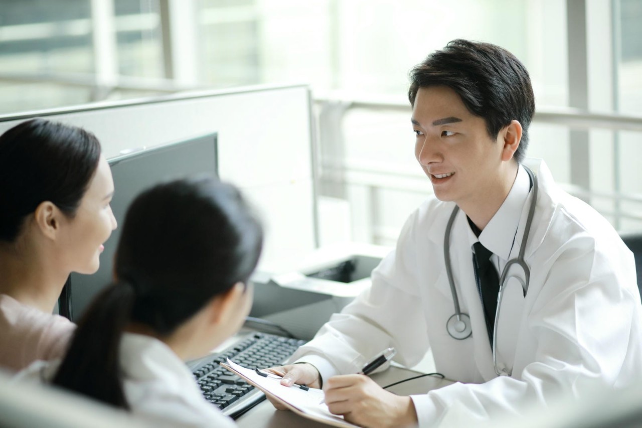 Smiling male doctor in a white coat with a stethoscope talking to two patients at a desk in a modern clinic.