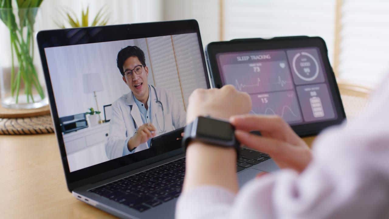 Telehealth consultation on a laptop while checking a smartwatch and health data on a tablet.