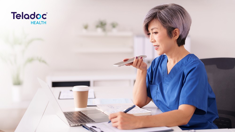 A nurse in blue scrubs speaks into her phone while writing in a notebook beside an open laptop in a bright office.